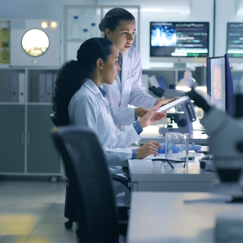 Two female scientists in lab coats focused on a computer screen, conducting research in a laboratory.